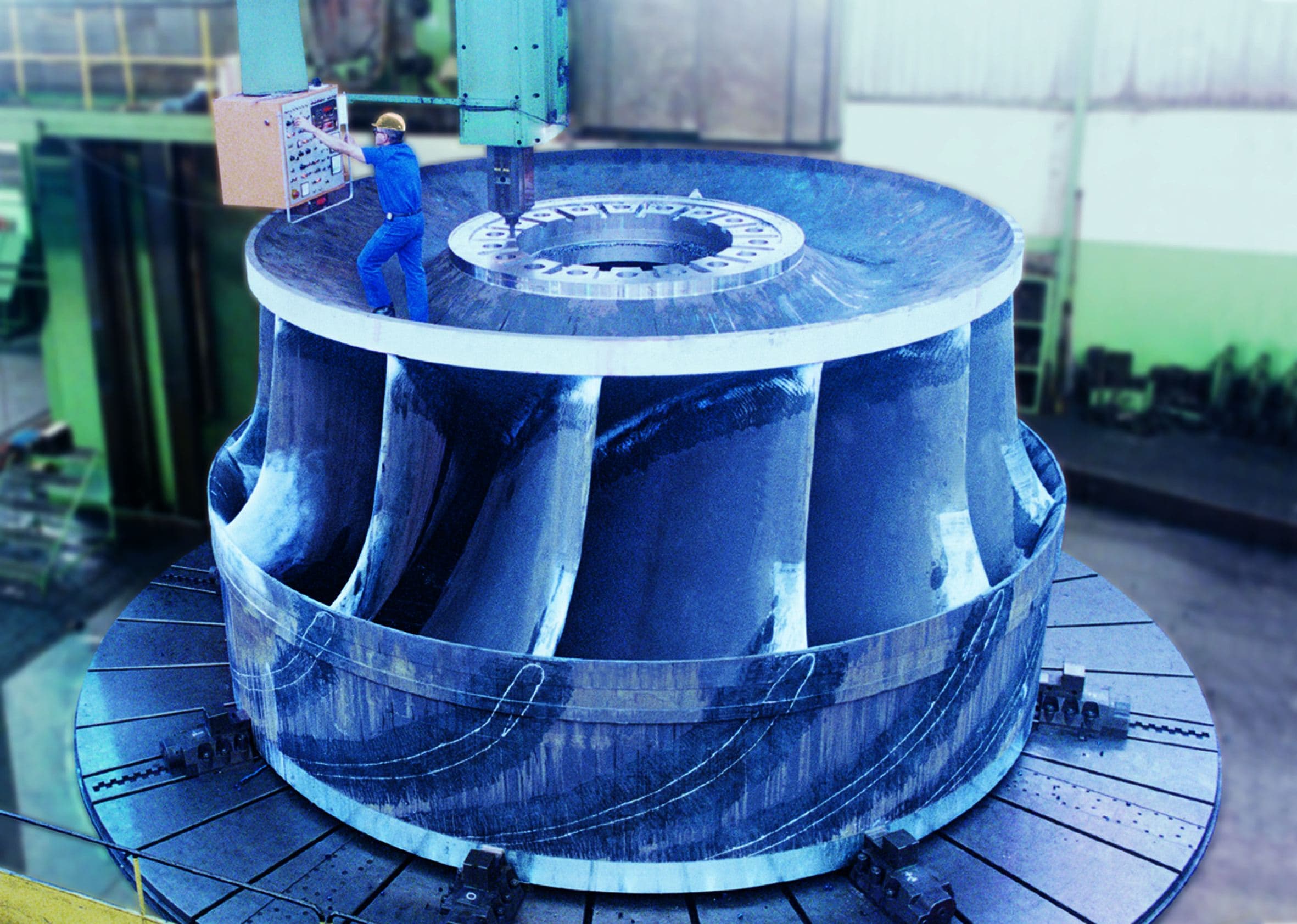 A Francis turbine runner mounted on a vertical carousel lathe in the workshop, a technician alongside it for scale.